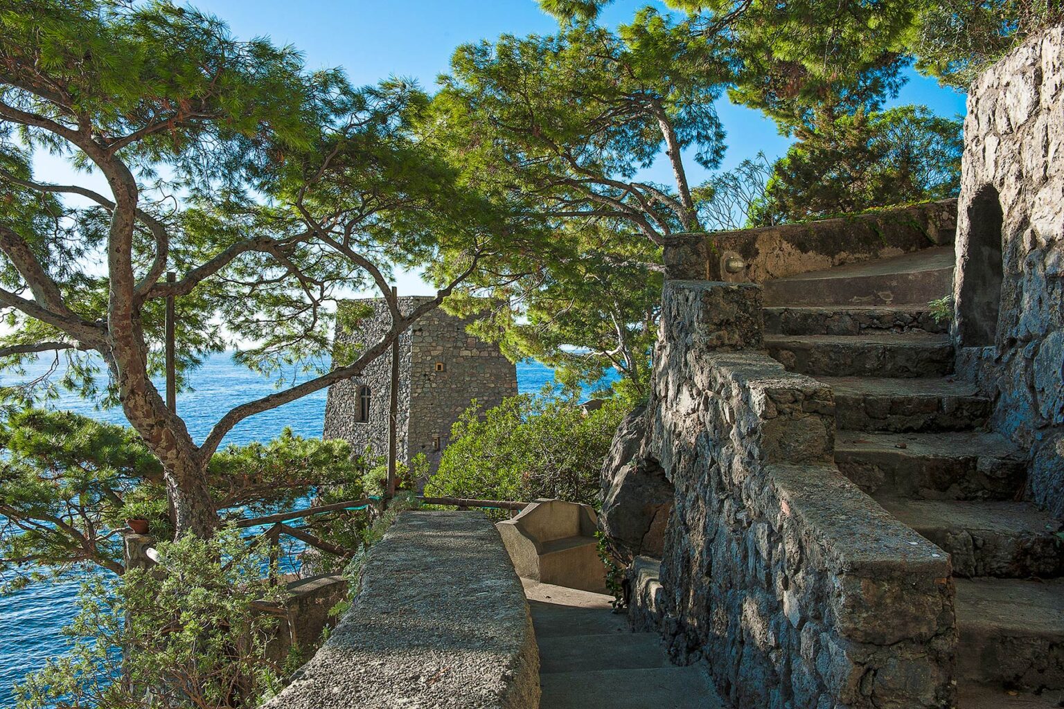 Stone staircase leading to a coastal lookout tower, surrounded by lush greenery and overlooking a serene blue sea, highlighting scenic natural beauty and historical architecture. | Only on SopranoVillas