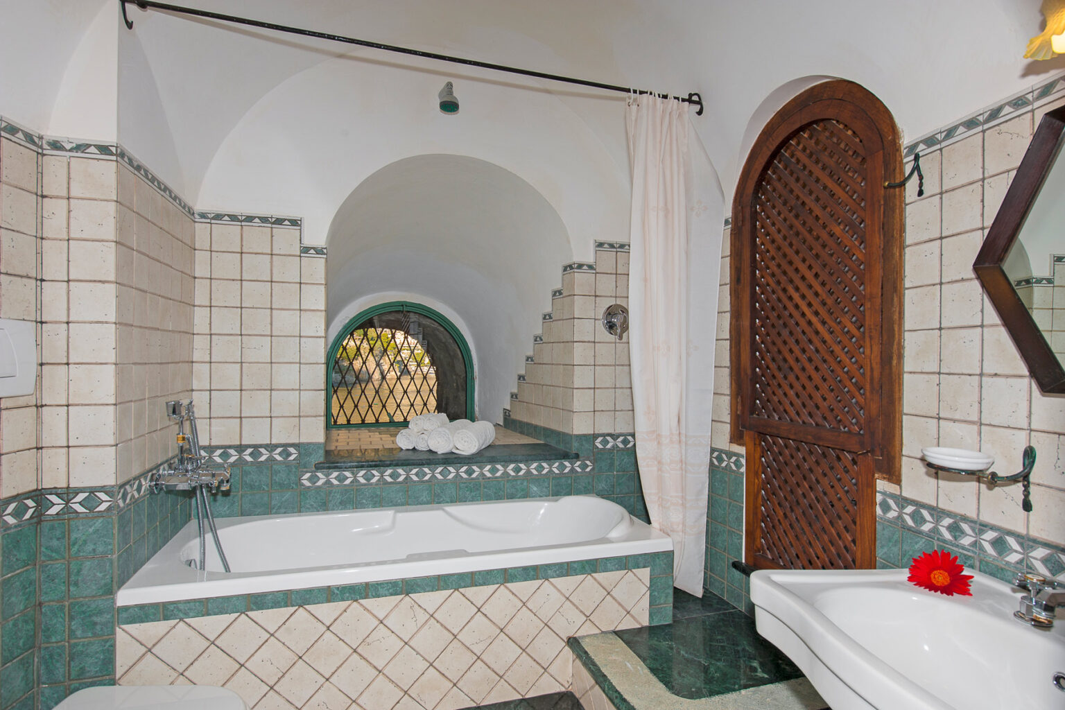 Elegant bathroom interior featuring a spacious bathtub, intricate tile work, and a wooden door. Natural light filters through decorative arches, creating a serene atmosphere. Ideal for relaxation and luxury home design inspiration. | Only on SopranoVillas