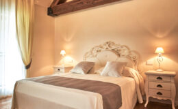 Elegant bedroom featuring a cozy double bed with decorative pillows, wooden beams on the ceiling, and soft lighting. The room includes stylish nightstands and sheer curtains, creating a warm and inviting atmosphere. Ideal for relaxation and comfort.