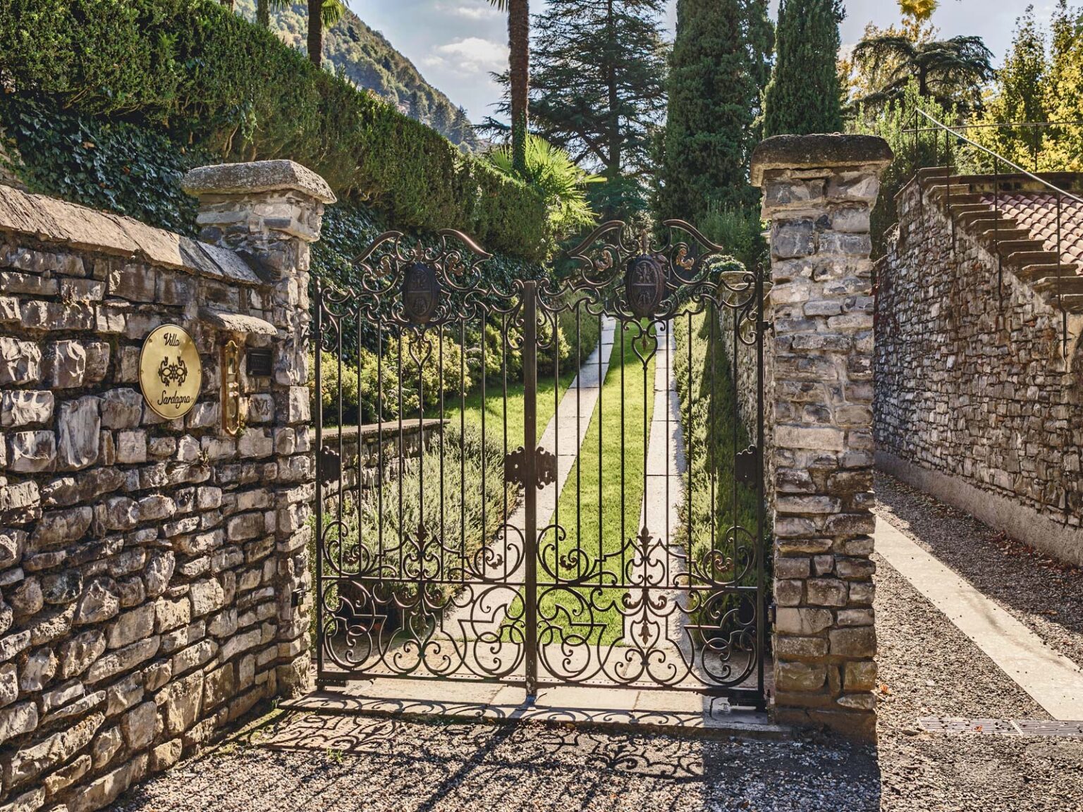 Wrought iron gate opening to a lush garden pathway, flanked by stone walls and greenery, ideal for showcasing outdoor landscaping design. | Only on SopranoVillas