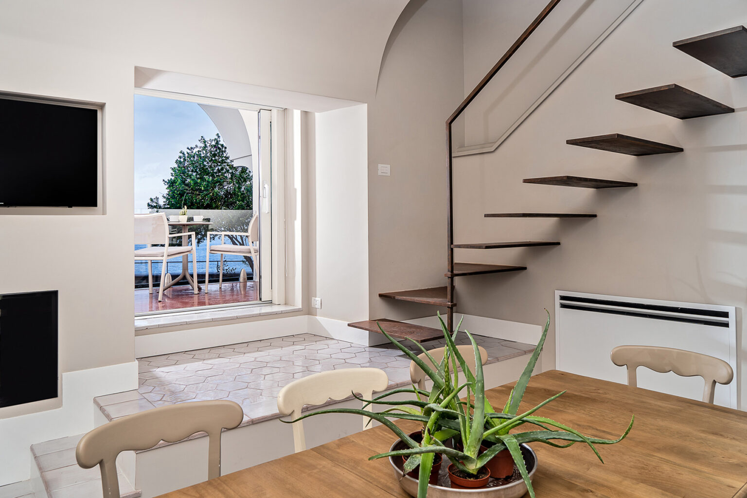 Modern interior of a stylish home featuring a staircase, dining area with a plant, and large windows overlooking a balcony with outdoor seating. | Only on SopranoVillas