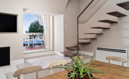 Modern interior of a stylish home featuring a staircase, dining area with a plant, and large windows overlooking a balcony with outdoor seating.