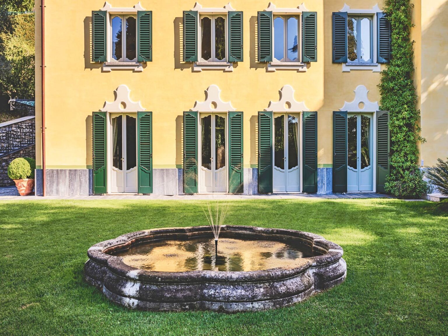 Elegant villa with yellow exterior and green shutters, featuring a circular fountain in the foreground and manicured lawn. | Only on SopranoVillas