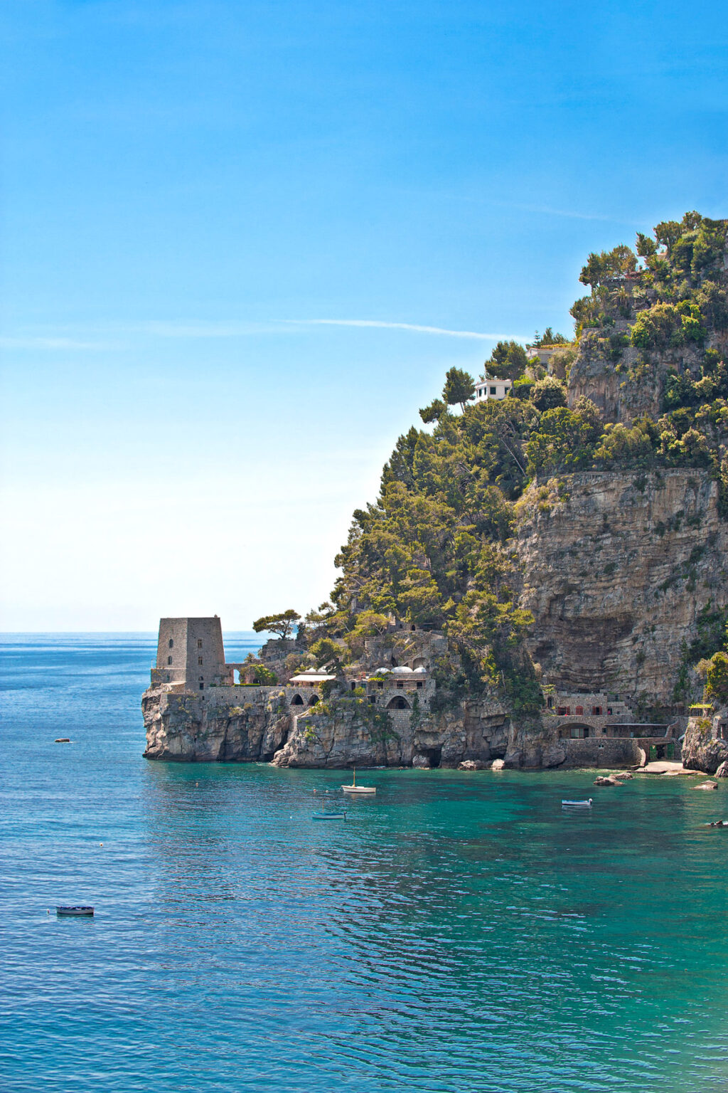 Scenic view of a coastal cliff in Italy featuring a historic watchtower and lush greenery overlooking the tranquil blue waters of the Mediterranean Sea. | Only on SopranoVillas