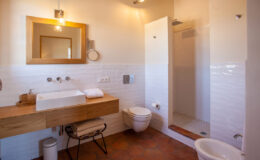 Modern bathroom interior featuring a wooden vanity with a sink, a large mirror, a shower stall, a toilet, and stylish lighting. The design combines functionality with a clean aesthetic, showcasing white tiles and warm tones.