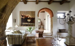 Modern living room interior featuring a striped sofa, wooden dining table, and a large artwork on the wall. The space includes rustic elements like exposed beams and an arched doorway, complemented by elegant decor such as a chandelier and decorative plants.