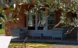 Cozy outdoor seating area with wicker furniture, framed by olive tree branches, next to a charming brick house. Ideal for relaxation and enjoying nature.