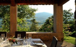 Elegant outdoor dining setup with a scenic view of mountains and greenery, featuring a beautifully arranged table with glassware and floral centerpiece under a rustic pergola.