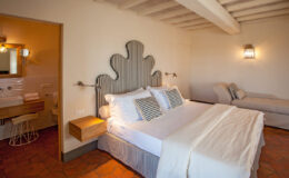 Spacious and stylish hotel bedroom featuring a king-sized bed with decorative pillows, a cozy seating area, and modern amenities. Natural light filters in through the windows, highlighting the elegant decor and warm color palette.