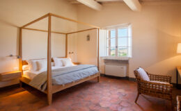 Spacious and modern bedroom featuring a wooden four-poster bed, natural light from large windows, terracotta flooring, and a cozy seating area with a wicker chair. Ideal for a relaxing getaway.