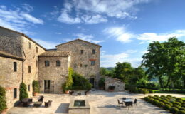 Historic stone villa surrounded by lush greenery, featuring a spacious outdoor patio with seating and a tranquil water feature under a bright blue sky.