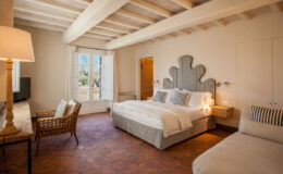 Spacious and elegantly designed bedroom featuring a king-sized bed with decorative headboard, cozy seating area, and large windows allowing natural light. The room showcases rustic tile flooring and stylish furnishings, creating a warm and inviting atmosphere.