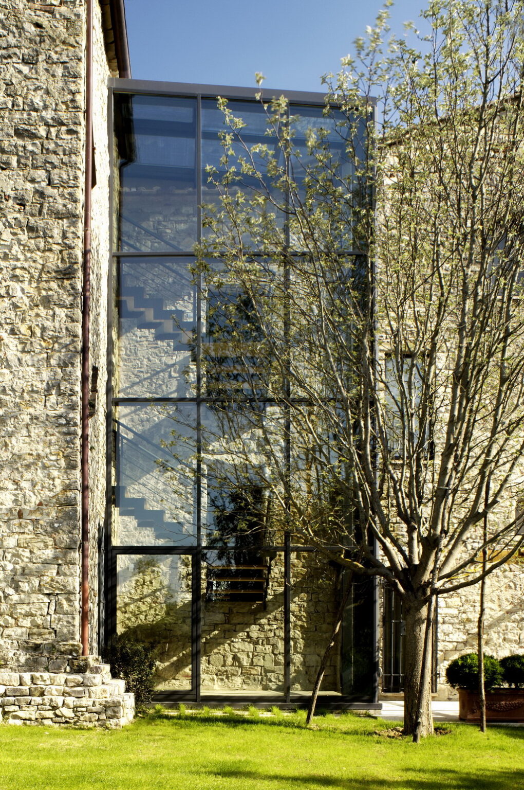 Modern glass staircase visible through a textured stone building facade, framed by a leafy tree. | Only on SopranoVillas