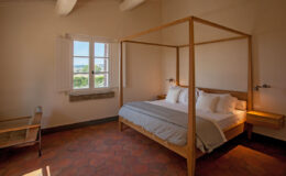Cozy bedroom featuring a wooden four-poster bed with white linens, natural light streaming through large windows, and a minimalist design with terracotta flooring and simple furnishings. Ideal for a relaxing getaway.