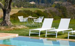 Relaxing outdoor space featuring white lounge chairs by a swimming pool, with a table and chairs set in a lush garden, surrounded by scenic views.