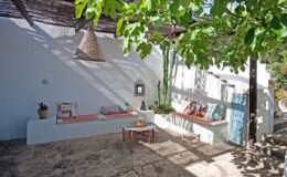 Cozy outdoor living space featuring white walls, rustic benches with cushions, and lush greenery, ideal for relaxation and entertaining in a tranquil setting.