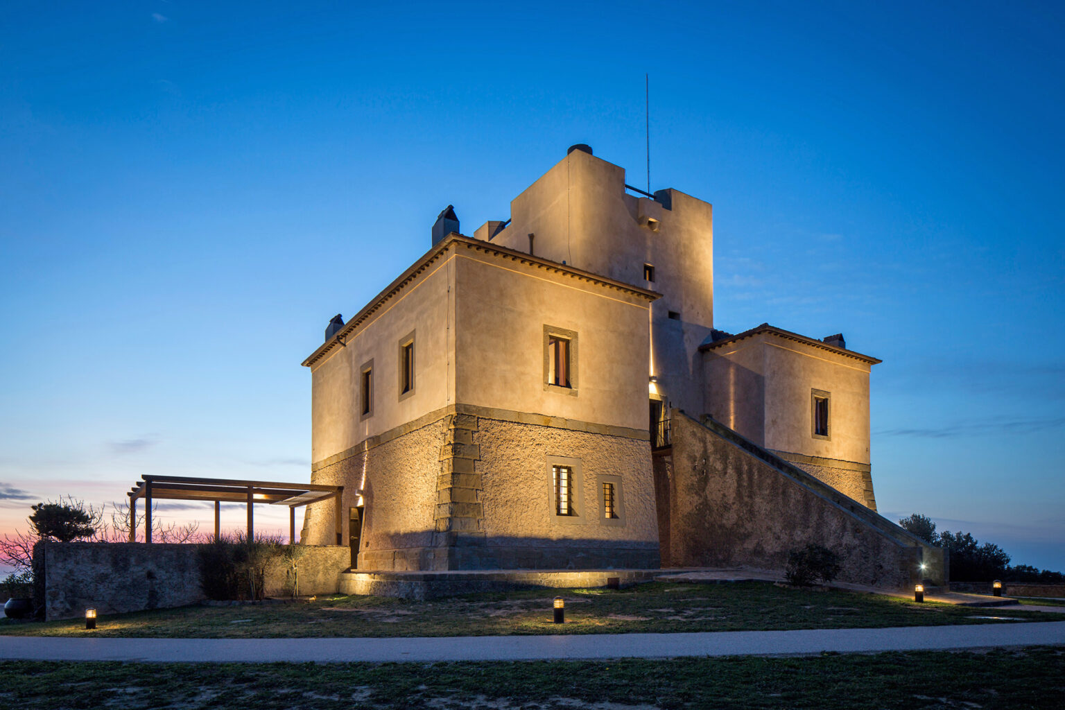 Historic stone castle illuminated at dusk, showcasing its architectural features against a twilight sky. The surrounding landscape includes a pathway and outdoor seating area, enhancing its scenic appeal. | Only on SopranoVillas