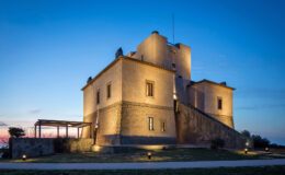 Historic stone castle illuminated at dusk, showcasing its architectural features against a twilight sky. The surrounding landscape includes a pathway and outdoor seating area, enhancing its scenic appeal.