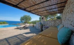 Lounge area with comfortable seating under a thatched roof, overlooking a serene swimming pool and the ocean, surrounded by lush greenery. Perfect for relaxation and enjoying coastal views.