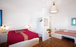 Cozy interior of a modern bedroom featuring a large bed with a red and beige blanket, a comfortable seating area with decorative cushions, and minimalist decor in a bright, white space.