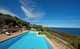 Scenic view of a luxurious swimming pool overlooking the ocean, surrounded by lush greenery and a clear blue sky, ideal for a relaxing getaway.
