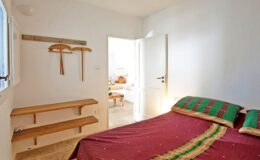 Cozy bedroom with a red and green patterned bedspread, featuring wooden shelves and a door leading to a bright, airy living space.