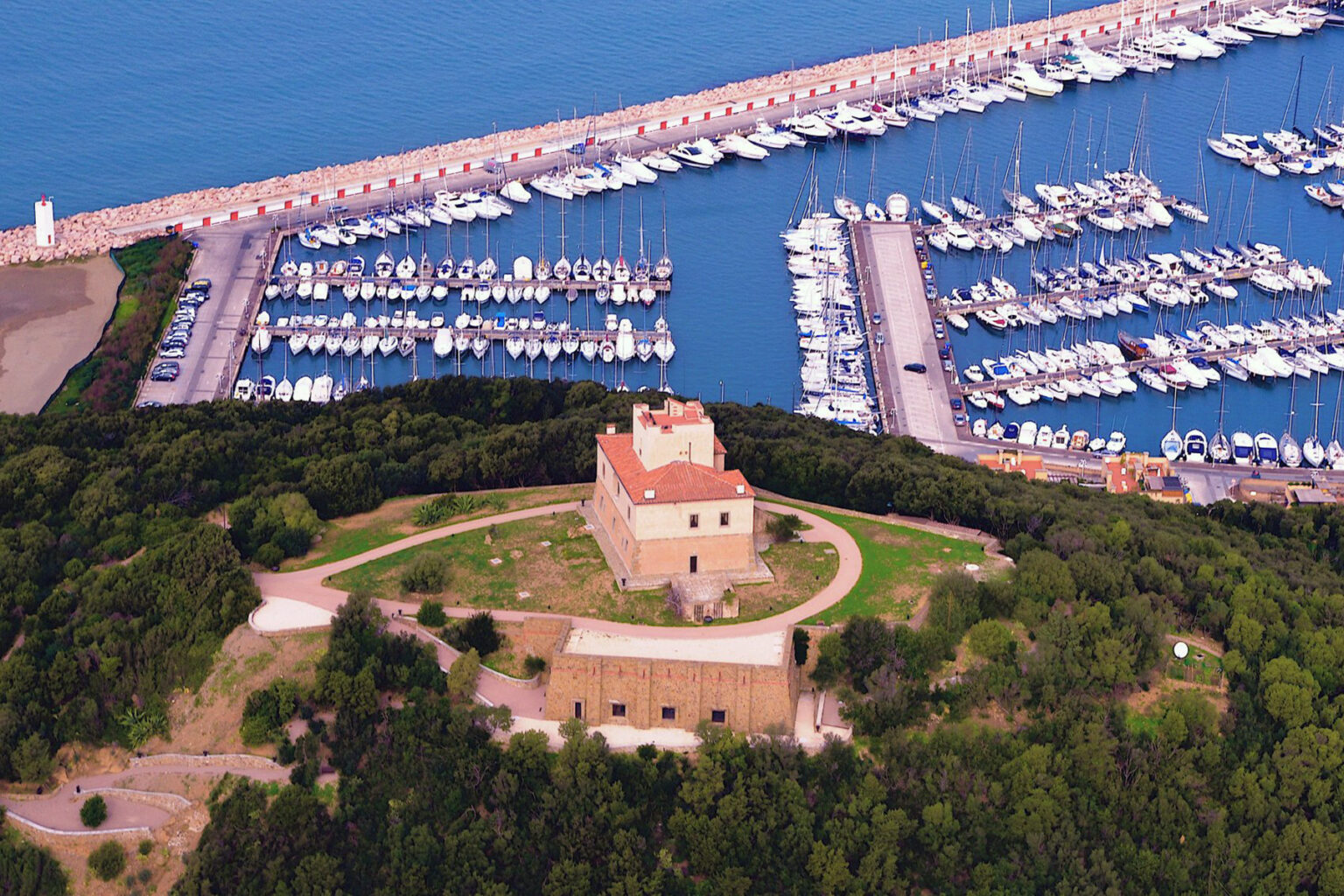 Aerial view of a historic building surrounded by lush greenery, overlooking a marina filled with yachts and boats, showcasing a picturesque coastal landscape. | Only on SopranoVillas