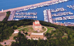 Aerial view of a historic building surrounded by lush greenery, overlooking a marina filled with yachts and boats, showcasing a picturesque coastal landscape.