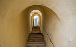 Narrow stone staircase leading through an arched hallway with warm yellow walls, illuminated by soft lighting, creating an inviting atmosphere.