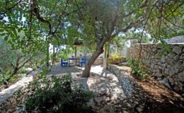 Scenic outdoor seating area surrounded by lush greenery, featuring a large tree and stone wall, ideal for relaxation and enjoying nature.
