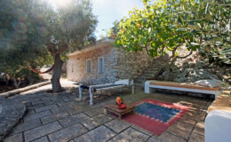Charming outdoor patio with a stone cottage, shaded by olive trees, featuring a wooden bench and colorful rug, perfect for relaxation in a serene garden setting.