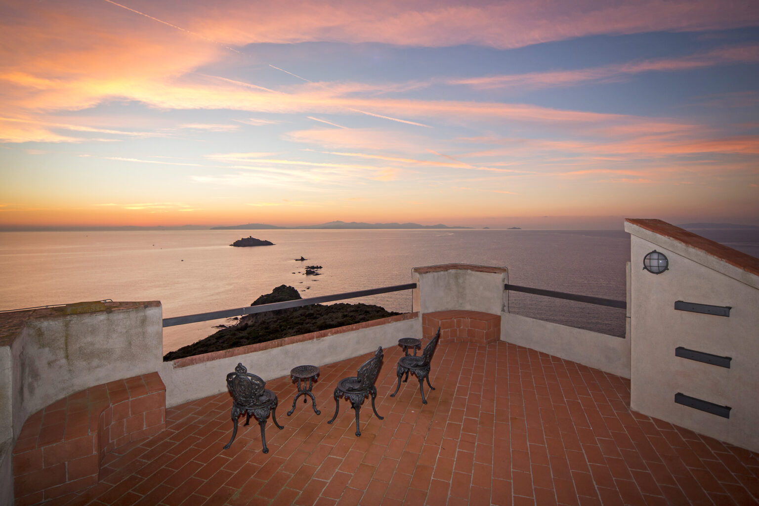 Scenic sunset view from a coastal terrace featuring four metal chairs and a table, overlooking calm waters and distant islands. The sky is painted with vibrant hues of orange, pink, and purple, creating a serene atmosphere. | Only on SopranoVillas