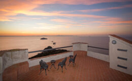 Scenic sunset view from a coastal terrace featuring four metal chairs and a table, overlooking calm waters and distant islands. The sky is painted with vibrant hues of orange, pink, and purple, creating a serene atmosphere.
