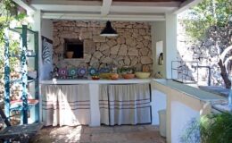 Outdoor kitchen with stone walls, wooden ceiling, and a variety of colorful dishes on the countertop, surrounded by greenery. Ideal for al fresco dining and cooking experiences.