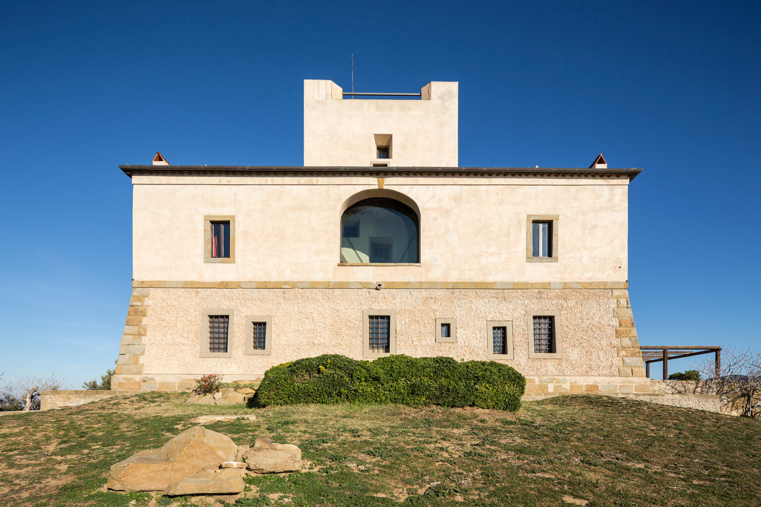 Historic stone building with a flat roof and large central window, set against a clear blue sky, surrounded by green grass and rocky terrain. | Only on SopranoVillas
