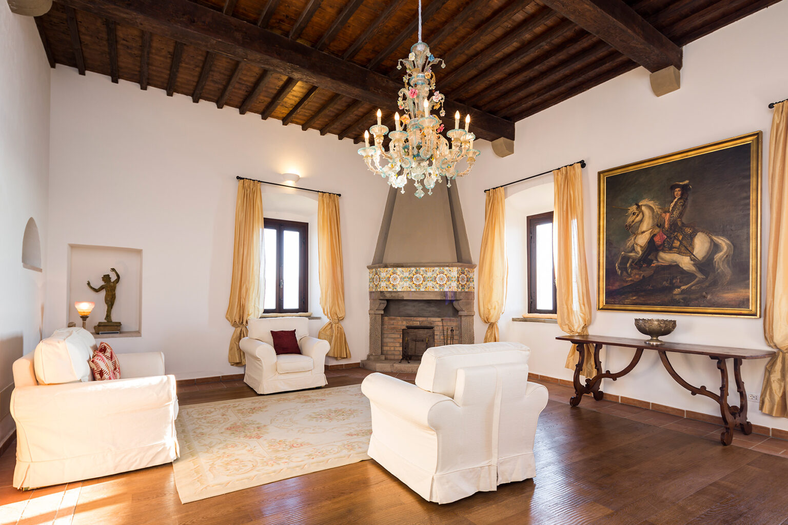 Spacious living room featuring white armchairs, a wooden ceiling, a chandelier, and a large painting of a horse. The room is adorned with elegant curtains and a decorative fireplace, creating a warm and inviting atmosphere. | Only on SopranoVillas