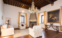 Spacious living room featuring white armchairs, a wooden ceiling, a chandelier, and a large painting of a horse. The room is adorned with elegant curtains and a decorative fireplace, creating a warm and inviting atmosphere.