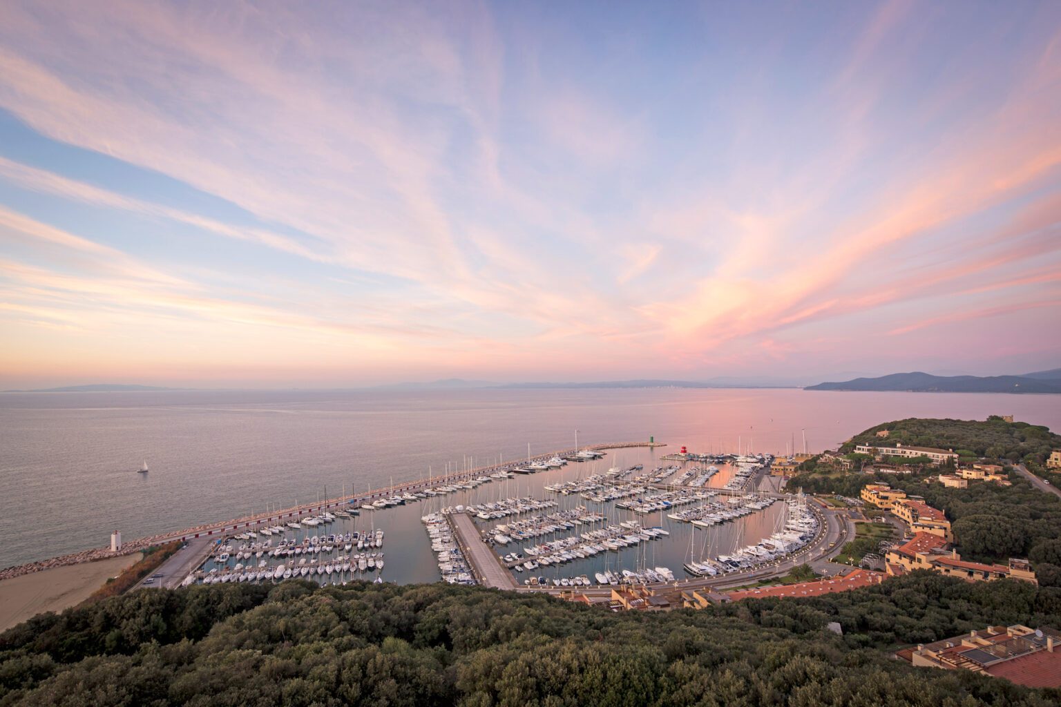 Aerial view of a marina at sunset, with numerous yachts docked along the shore, surrounded by lush greenery and a tranquil sea reflecting the colorful sky. | Only on SopranoVillas