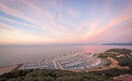 Aerial view of a marina at sunset, with numerous yachts docked along the shore, surrounded by lush greenery and a tranquil sea reflecting the colorful sky.