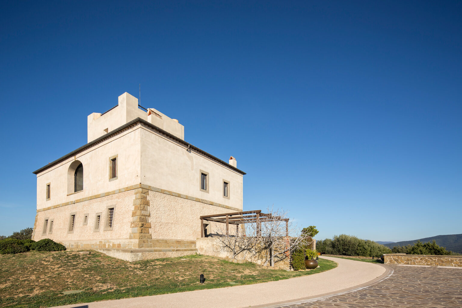 Historic stone building with a flat roof and large windows, surrounded by greenery and a paved path under a clear blue sky. Ideal for architectural and travel photography. | Only on SopranoVillas