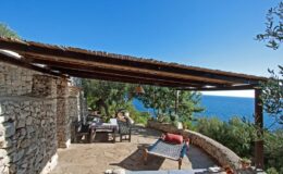 Cozy outdoor patio with a stone wall, wooden pergola, and a stunning view of the ocean, featuring a dining table and lounge chair under clear blue skies.