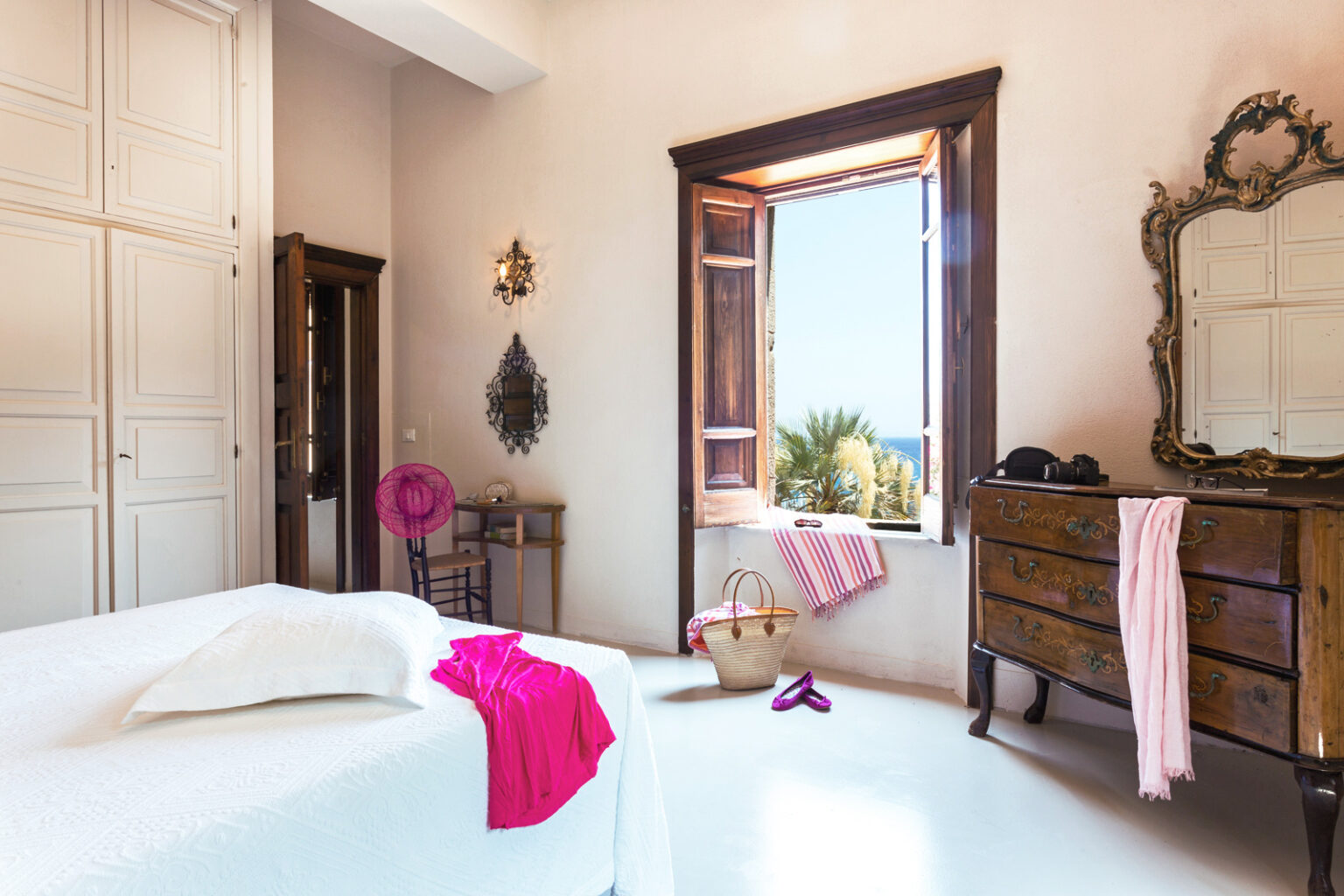 Bright and airy bedroom featuring a large window with scenic views, a wooden dresser, and white bedding accented with pink decor. Natural light enhances the space's charm, complemented by stylish accessories and a relaxed atmosphere. | Only on SopranoVillas