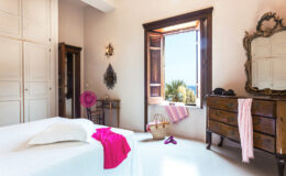 Bright and airy bedroom featuring a large window with scenic views, a wooden dresser, and white bedding accented with pink decor. Natural light enhances the space's charm, complemented by stylish accessories and a relaxed atmosphere.