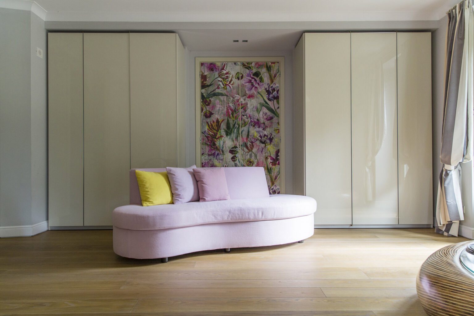 Stylish modern living room featuring a curved pink sofa with yellow accent pillows, complemented by sleek cabinetry and a vibrant floral accent wall. The warm wooden floor enhances the cozy atmosphere. | Only on SopranoVillas