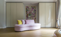 Stylish modern living room featuring a curved pink sofa with yellow accent pillows, complemented by sleek cabinetry and a vibrant floral accent wall. The warm wooden floor enhances the cozy atmosphere.