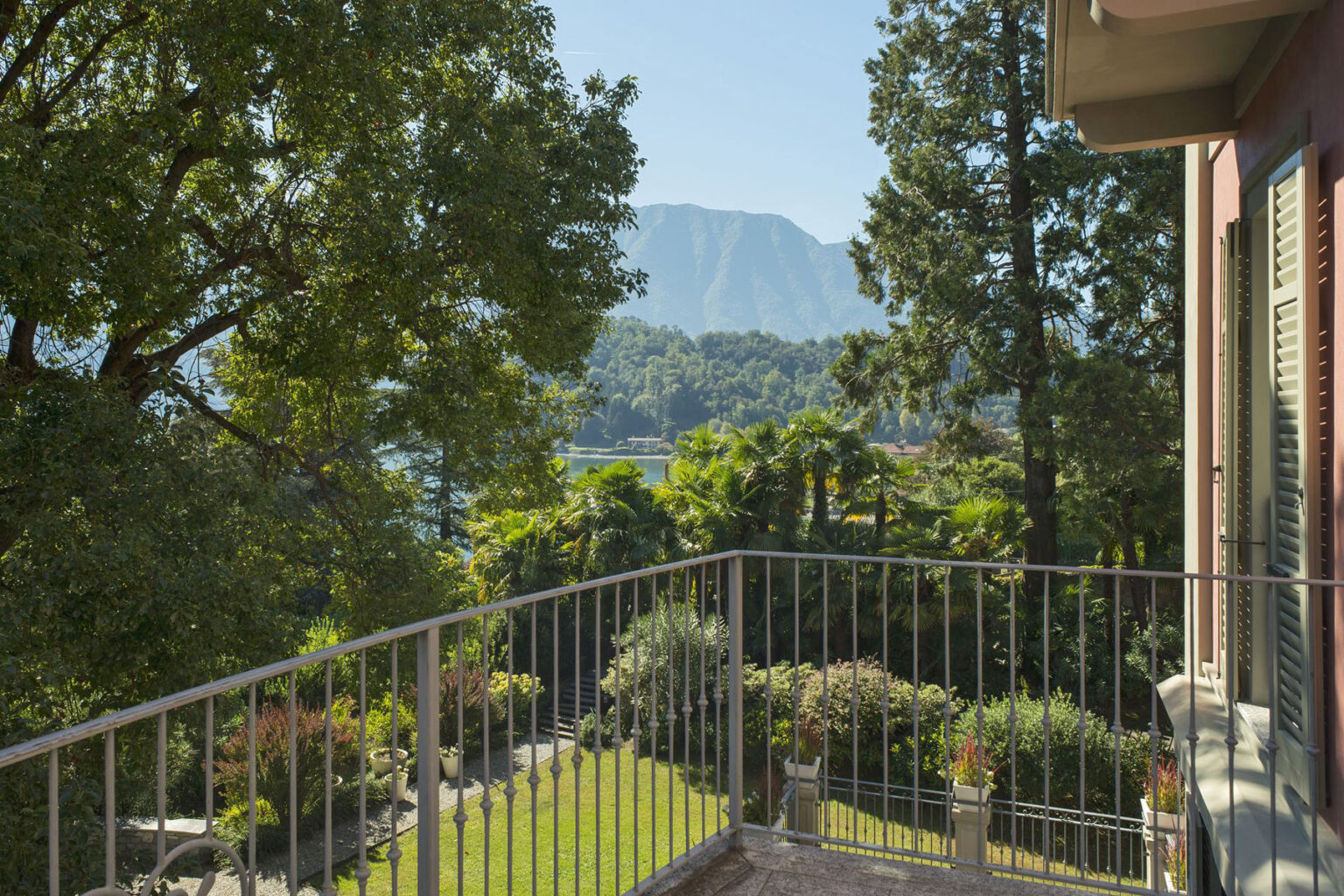 Scenic view from a balcony overlooking lush greenery and mountains, with a clear blue sky and vibrant foliage in the foreground. Ideal for showcasing serene outdoor living spaces and natural landscapes. | Only on SopranoVillas