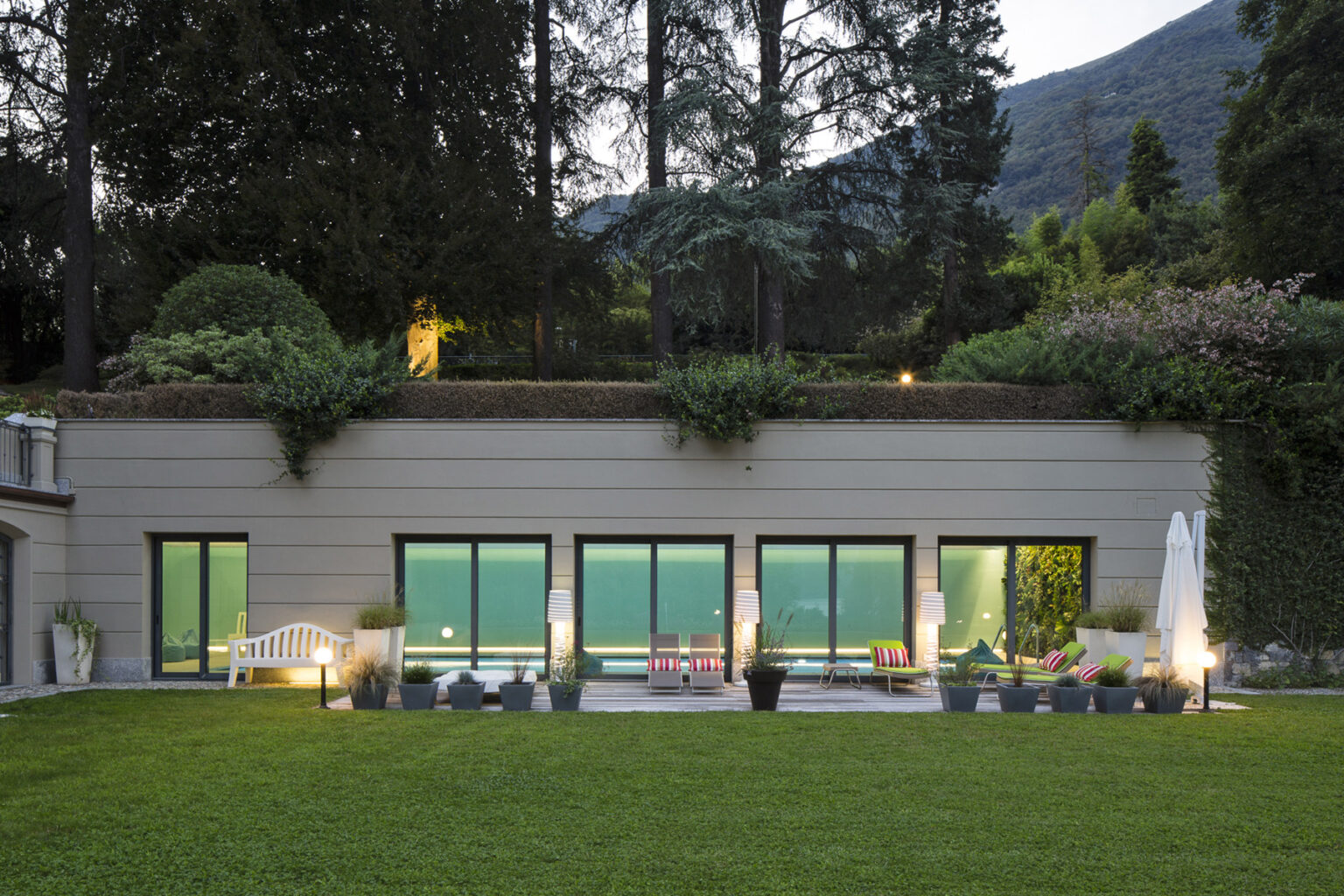 Modern hillside villa with large glass windows, surrounded by lush greenery and illuminated outdoor seating area, showcasing a serene landscape and contemporary architecture. | Only on SopranoVillas