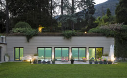 Modern hillside villa with large glass windows, surrounded by lush greenery and illuminated outdoor seating area, showcasing a serene landscape and contemporary architecture.