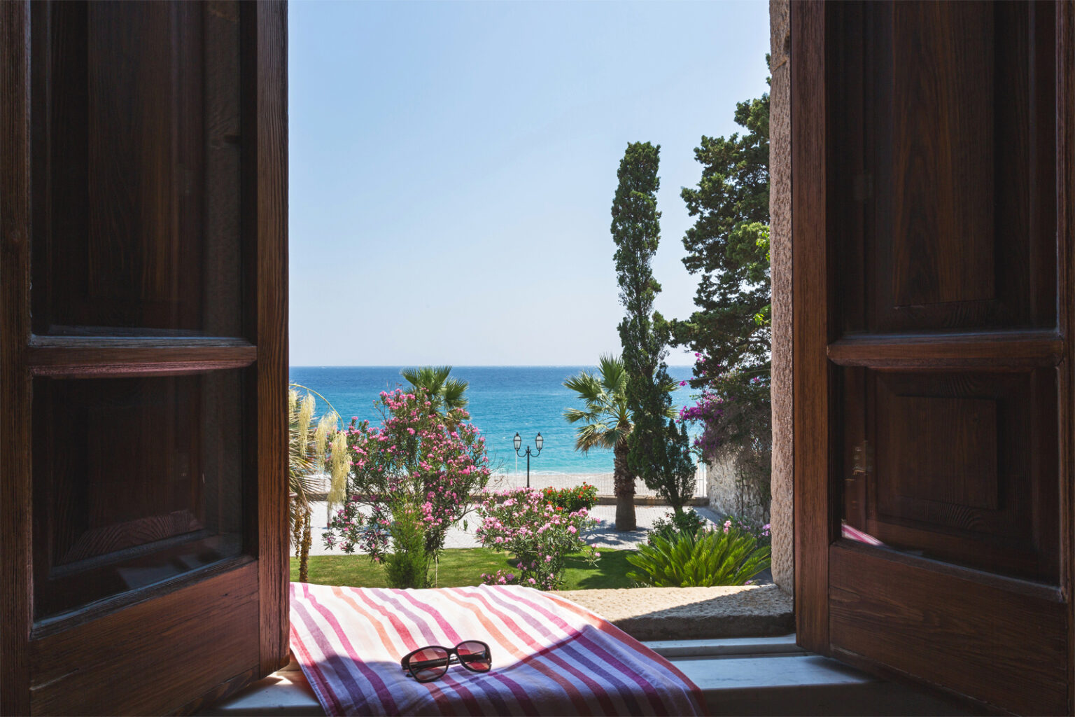 View from an open wooden window overlooking a serene beach with turquoise waters, palm trees, and blooming flowers, creating a tranquil coastal atmosphere. A pair of sunglasses rests on a striped towel in the foreground. | Only on SopranoVillas