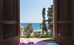 View from an open wooden window overlooking a serene beach with turquoise waters, palm trees, and blooming flowers, creating a tranquil coastal atmosphere. A pair of sunglasses rests on a striped towel in the foreground.
